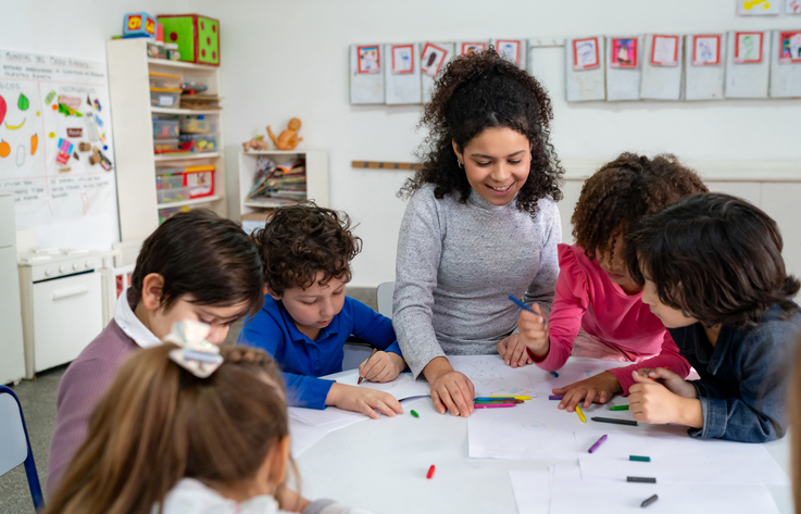 Focused school kids coloring with crayons while cheerful teacher supervises.