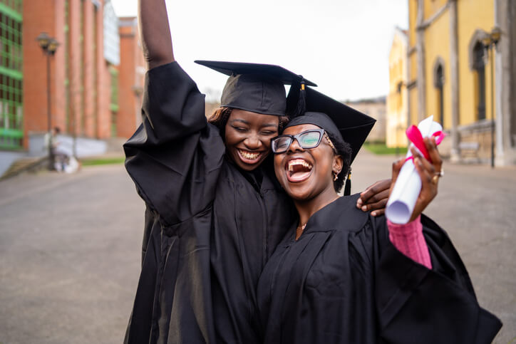 Two happy Black female graduates are celebrating their graduation by hugging and raising their arms in the air.