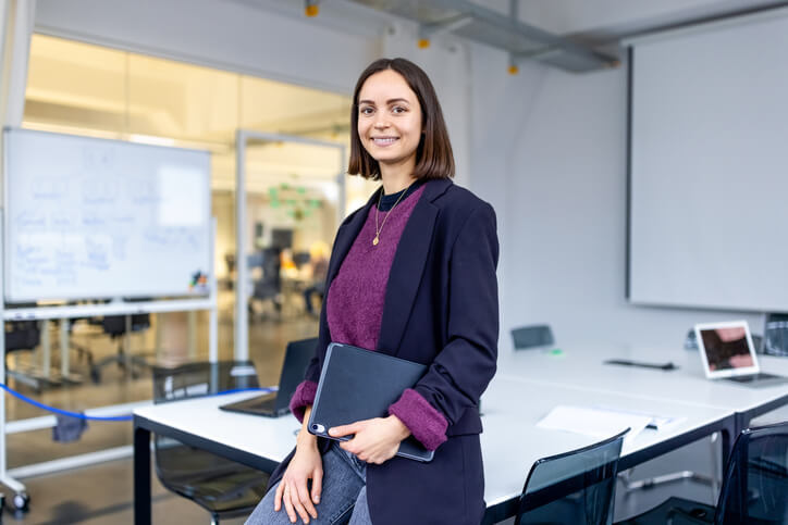 Portrait of a smiling businesswoman standing in an office meeting room. She is wearing a navy blue blazer and purple sweater, and is holding a tablet device.