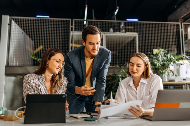 Three colleagues sitting at a desk in an office. They are looking at ideas on a laptop and and brainstorming marketing ideas.