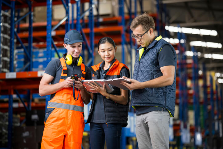A supply chain team of three having a strategy meeting in a distribution warehouse to determine the prioritization order of shipments.