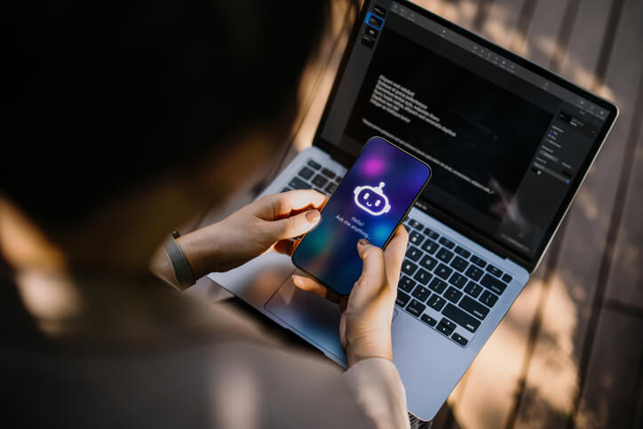 Closeup of a businesswoman using a generative AI app on her smartphone to assist her while working on her laptop in an office park.