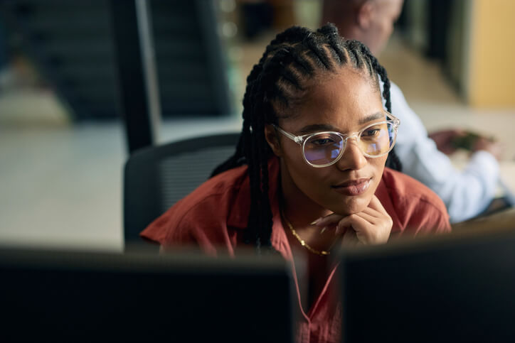 Black female front-end web developer focused on her work, coding on dual monitors in a modern office environment.