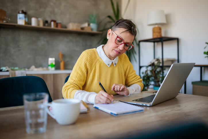 Woman in her forties with gray hair taking an online class at home. She is writing notes while looking at her laptop computer.