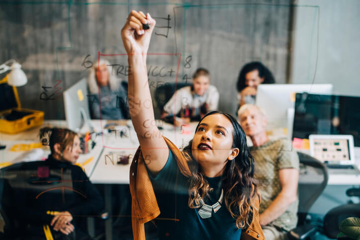 Businesswoman explaining her strategy to colleagues while writing on a glass marker board at an IT company.