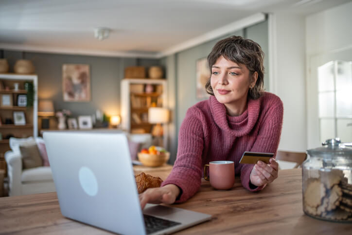 A woman in her forties sits at her kitchen counter and checks her monthly budget via online banking, using a digital tablet and credit card.