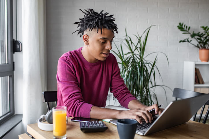 Black male college student working on his laptop at home. He has a calculator next to his laptop and is typing on the keyboard.