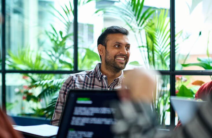 Cheerful South Asian male computer scientist sitting at a table in a meeting room. He is listening to his coworkers speaking during a business meeting.