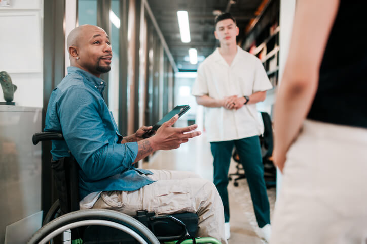 A budget analyst in a wheelchair chatting with two of his younger colleagues. He is holding a tablet device with a spreadsheet of financial data.