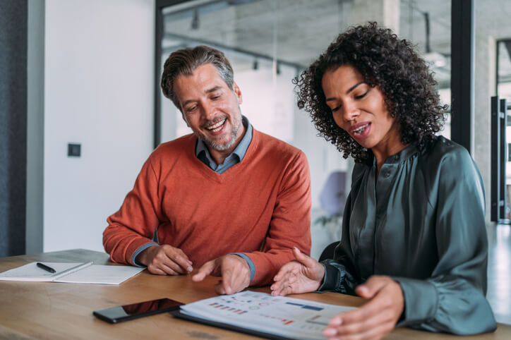 Two colleagues talking together while sitting in a modern office. They are working on business reports for a presentation.