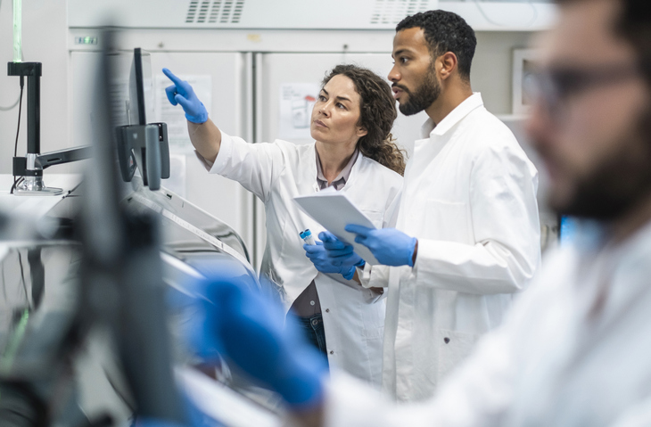 A mid-adult Caucasian female professor and a Black male college student, both dressed in lab coats, engage intently over research data in a bright laboratory setting.
