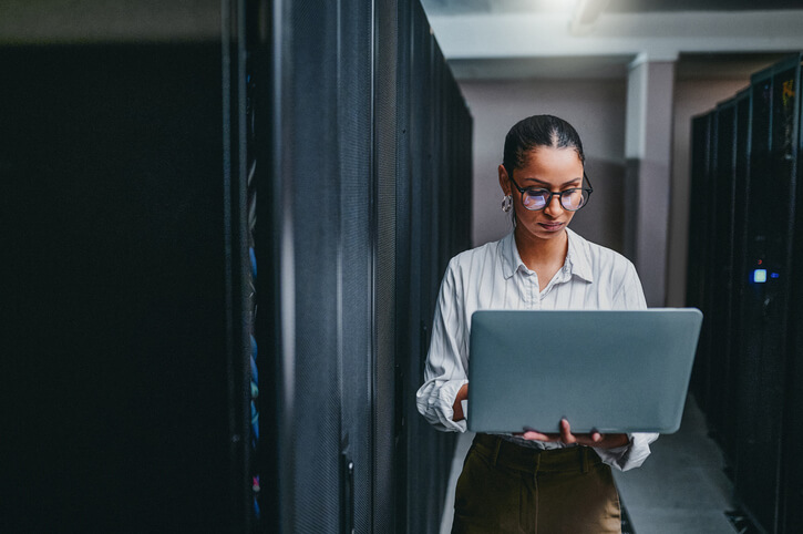 Black female cybersecurity professional using a laptop while working in a server room.
