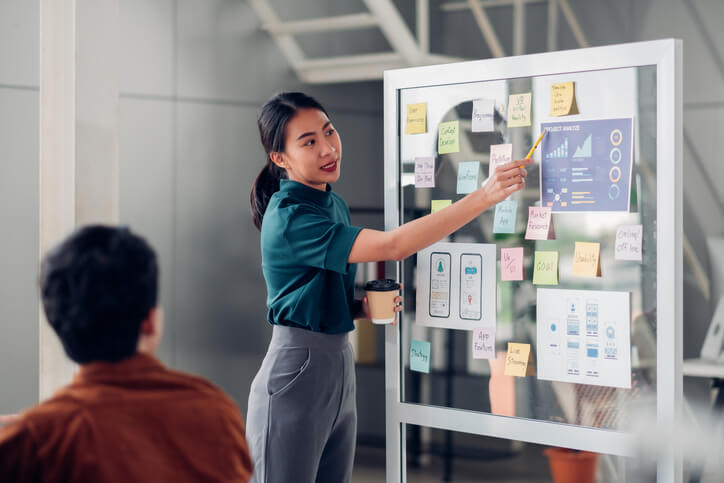 Female Asian UX designer presenting a mobile app interface design on a whiteboard during a meeting at the office.