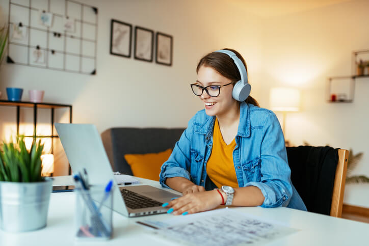 Joven sonriente con auriculares y gafas participando en un curso en línea de español desde casa. Está sentada en un escritorio con una computadora portátil, libros y material de oficina, mostrando interés y concentración durante la clase virtual.