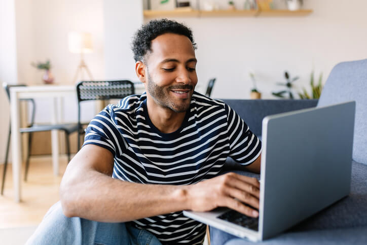 Black man using laptop computer while resting on sofa at home.