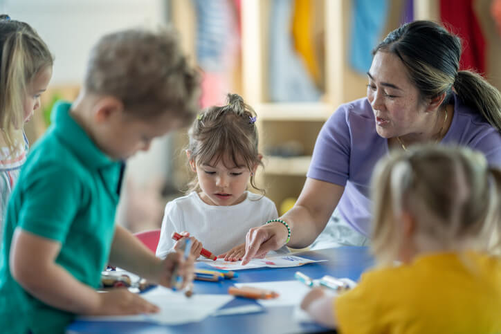 A female Asian American teacher assisting a group of preschool kids as they make arts and crafts at a table.