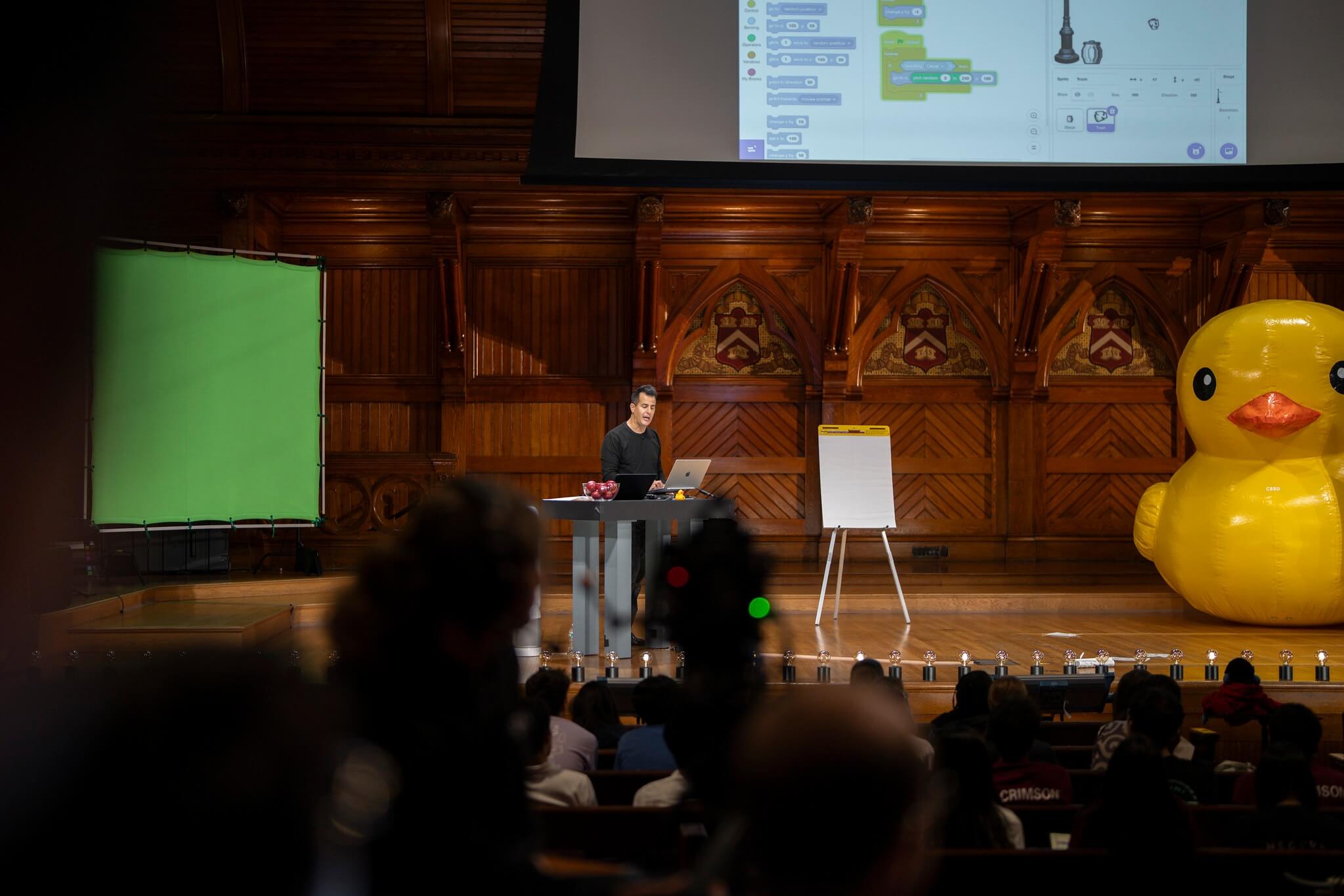 CS50 lecture in Sanders Theatre with Professor David J. Malan, featuring the course’s signature yellow rubber duck mascot.